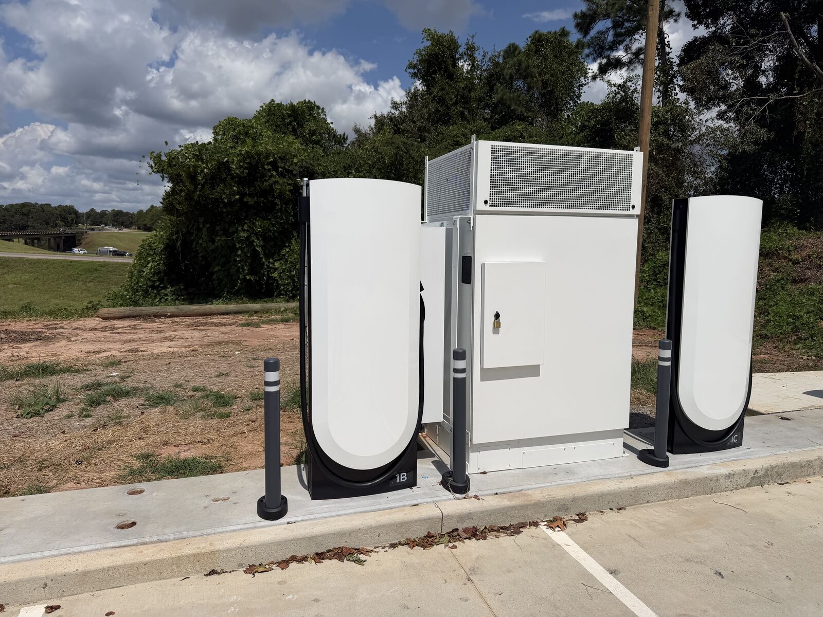 Two Tesla V4 Supercharger posts labeled 1B and 1C flanking a single equipment cabinet on a new concrete pad